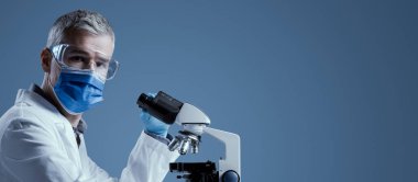 Tıp bilimci yüz maskesi takıyor ve laboratuvarda mikroskopla çalışıyor. Kameraya bakıyor.