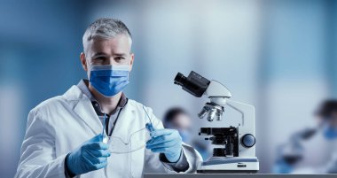 Tıp bilimci yüz maskesi takıyor ve laboratuvarda mikroskopla çalışıyor. Kameraya bakıyor.