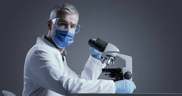 Medical scientist wearing a face mask and working in the laboratory with a microscope, he is looking at camera