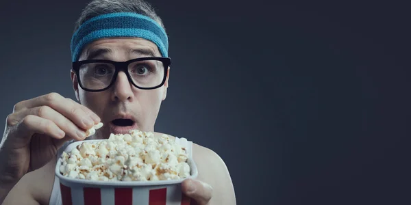 Funny guy with glasses and headband watching movies, he is staring at the screen with mouth open and eating popcorn