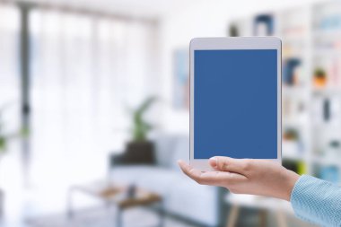 Elinde boş ekran ve evin içi olan bir tablet tutan kadın, akıllı ev otomasyonu kavramı.