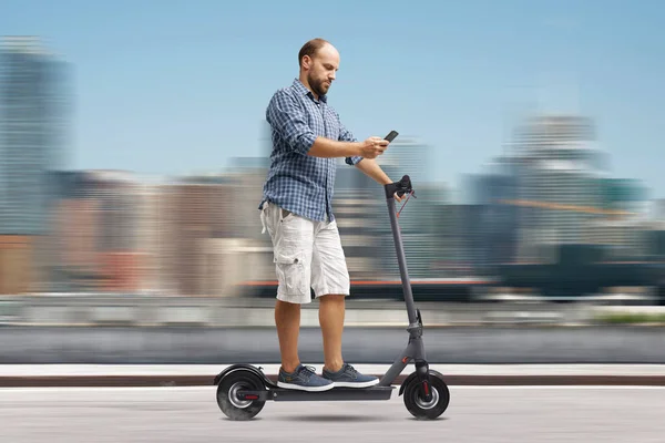 Careless man using a smartphone while riding an electric scooter, city in the background, sustainable mobility concept