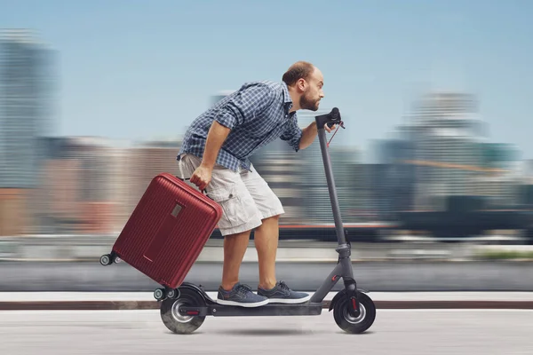 Traveller man holding a trolley bag and riding a fast electric scooter, city in the background, sustainable mobility concept