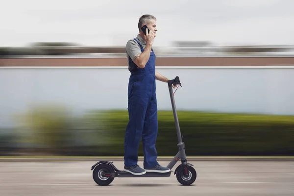 Express repair service: repairman having a phone call with his smartphone and riding a fast electric scooter, city in the background