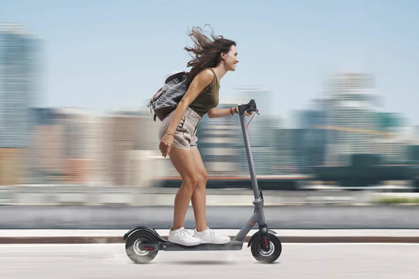 電動スクーターに乗っている幸せな屈託のない若い女性 背景の街並み — ストック写真