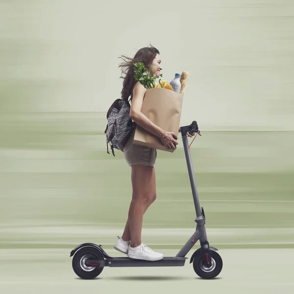 Young woman holding a paper bag with groceries and riding a fast electric scooter, sustainable mobility concept