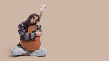 Şık hippi kadın bacak bacak üstüne atmış, elinde akustik gitar, müzik ve ilham konseptiyle oturuyor.