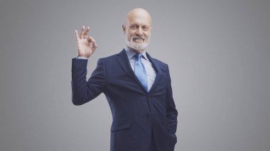 Gülümseyen şirket işadamı iyi bir jest yapıyor ve gri arka planda izole edilmiş kameraya bakıyor.