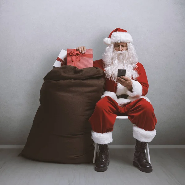 厌倦了的圣诞老人坐在那里 一边用智能手机聊天 一边提着满满一袋礼物 — 图库照片