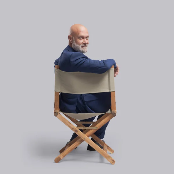Confident film director sitting on the director\'s chair and looking at camera: video production, filmmaking and film industry concept