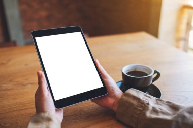 Siyah tabletli, beyaz ekranlı ve ahşap masada kahve fincanı olan ellerin görüntüsü. 