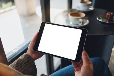 Kafedeki boş beyaz masaüstü ekran ile siyah tablet bilgisayarını tutan bir kadının görüntüsü.