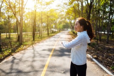 Genç Asyalı bir kadın şehir parkında koşmadan önce kollarını esnetiyor. 