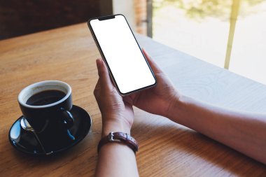 Elinde siyah cep telefonu olan, boş ekranlı, ahşap sehpada kahve fincanı olan bir kadının görüntüsü.