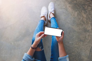 Üst görünümde, yerde otururken elinde beyaz ekranlı siyah bir cep telefonu tutan bir kadının görüntüsü var.