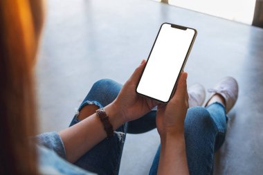 Yerde otururken elinde beyaz ekranlı siyah bir cep telefonu tutan bir kadının görüntüsü.