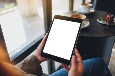 Kafedeki boş beyaz masaüstü ekran ile siyah tablet bilgisayarını tutan bir kadının görüntüsü.