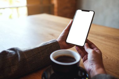 Ellerin ahşap masada kahve fincanı olan boş ekranlı cep telefonunu tutarken görüntüsü.