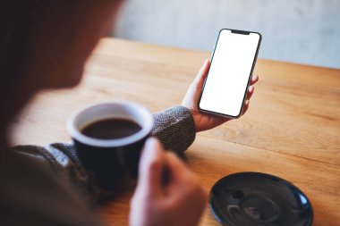 Ahşap masada kahve içerken elinde boş beyaz ekranla cep telefonu tutan bir kadının görüntüsü.