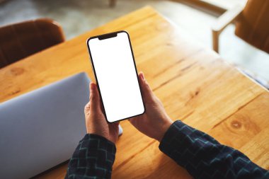 Masasında dizüstü bilgisayarı olan, boş beyaz masaüstü ekranlı cep telefonu tutan bir kadının görüntüsü. 