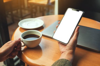 Kahve içerken elinde boş beyaz masaüstü ekranlı cep telefonu tutan bir kadının görüntüsü. 