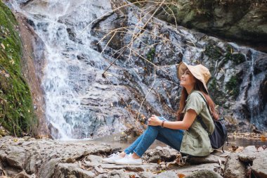 Güzel bir Asyalı kadın şelalenin önündeki kayanın üzerinde oturuyor.