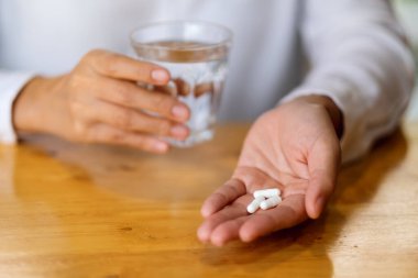 Beyaz haplar ve bir bardak su tutan bir kadının yakın plan görüntüsü.