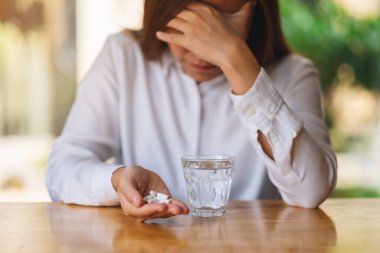 Baş ağrısı çeken bir kadının, elinde beyaz bir hap ve masada bir bardak su tutarken ki görüntüsü.