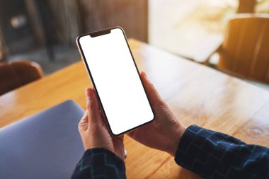 Masasında dizüstü bilgisayarı olan, boş beyaz masaüstü ekranlı cep telefonu tutan bir kadının görüntüsü. 