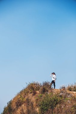 Mavi arka planda dağların tepesinde yürüyüş yapan ve ayakta duran bir kadın.