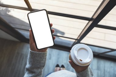 Üst görünümde, elinde siyah bir cep telefonu olan bir kadının kahve fincanı olan beyaz ekranlı görüntüsü var.