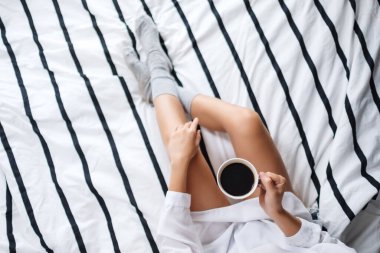 Sabahları beyaz bir yatakta elinde sıcak kahve tutan ve içen bir kadının görüntüsü.