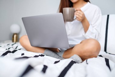 Bilgisayarda çalışan, beyaz bir yatakta oturup kahve içen ev konseptiyle uğraşan bir kadının yakın plan görüntüsü.