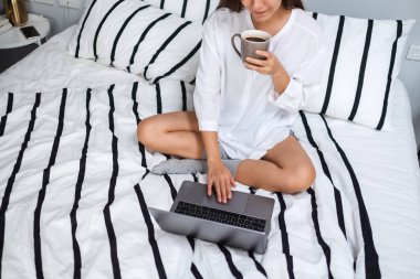 Güzel bir kadının dizüstü bilgisayarı kullanırken ve çalışırken çekilmiş resmi. Evdeki rahat beyaz bir yatakta oturup kahve içerken.
