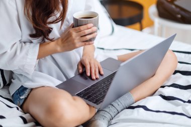 Laptop bilgisayar kullanan ve çalışan bir kadının yakın plan görüntüsü. Evdeki rahat beyaz bir yatakta oturup kahve içen bir kadın.