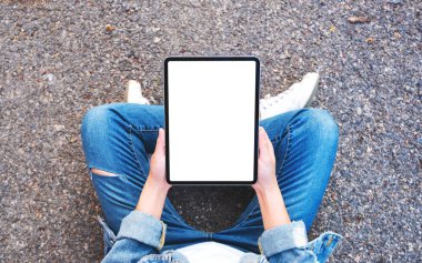 Üst görüntüde, elinde siyah tablet olan beyaz bir kadın açık havada otururken görüntüsü var. 