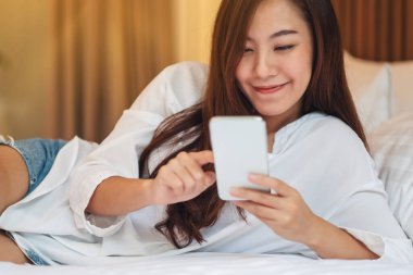 Güzel Asyalı bir kadının beyaz bir yatakta uzanmış cep telefonunu kullanırken ve ona bakarken yakın plan görüntüsü.