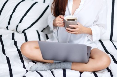 Laptop bilgisayar kullanan ve çalışan bir kadının yakın plan görüntüsü. Evdeki rahat beyaz bir yatakta oturup kahve içen bir kadın.