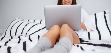 Evdeki rahat beyaz bir yatakta otururken dizüstü bilgisayar kullanan ve çalışan bir kadının...