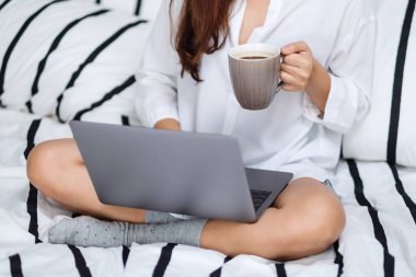 Laptop bilgisayar kullanan ve çalışan bir kadının yakın plan görüntüsü. Evdeki rahat beyaz bir yatakta oturup kahve içen bir kadın.