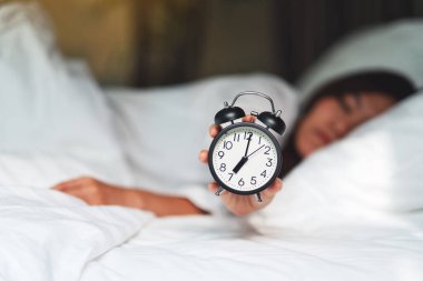 Asyalı bir kadın sabah beyaz bir yatakta uyurken çalar saati tutuyor ve gösteriyor.