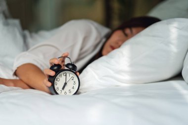 Asyalı bir kadın sabahın köründe beyaz bir yatakta uyurken çalar saati kapatıyor.