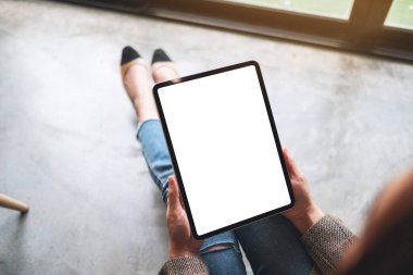 Yerde otururken boş beyaz masaüstü ekranlı tablet bilgisayarını tutan ve kullanan bir kadının en üst görünüm görüntüsü 