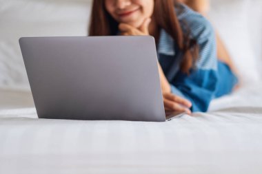 Güzel bir Asyalı kadın evdeki rahat beyaz bir yatakta yatarken dizüstü bilgisayar kullanıyor ve çalışıyor.