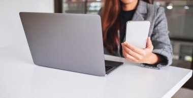 Genç ve güzel bir iş kadını cep telefonunu ahşap masada dizüstü bilgisayarla tutuyor ve kullanıyor.
