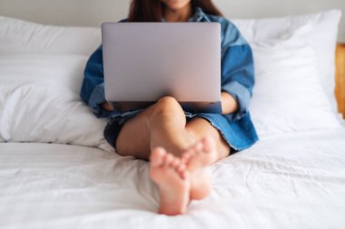 Güzel bir Asyalı kadın evdeki rahat beyaz bir yatakta yatarken dizüstü bilgisayar kullanıyor ve çalışıyor.