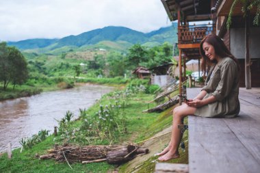 Dağlar ve doğa geçmişiyle nehir kenarında otururken cep telefonu kullanan güzel bir Asyalı kadın.
