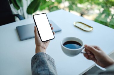 Kahve içerken elinde boş beyaz masaüstü ekranıyla cep telefonu tutan bir kadının görüntüsü 