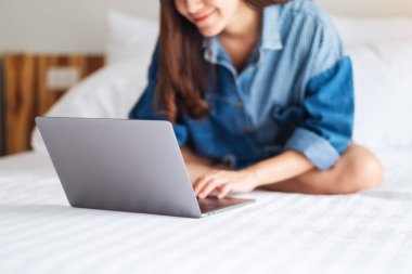 Güzel bir Asyalı kadın evdeki rahat beyaz bir yatakta otururken dizüstü bilgisayar kullanıyor ve çalışıyor.