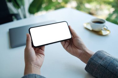 Masasında dizüstü bilgisayarı olan, boş beyaz masaüstü ekranlı cep telefonu tutan bir kadının görüntüsü. 